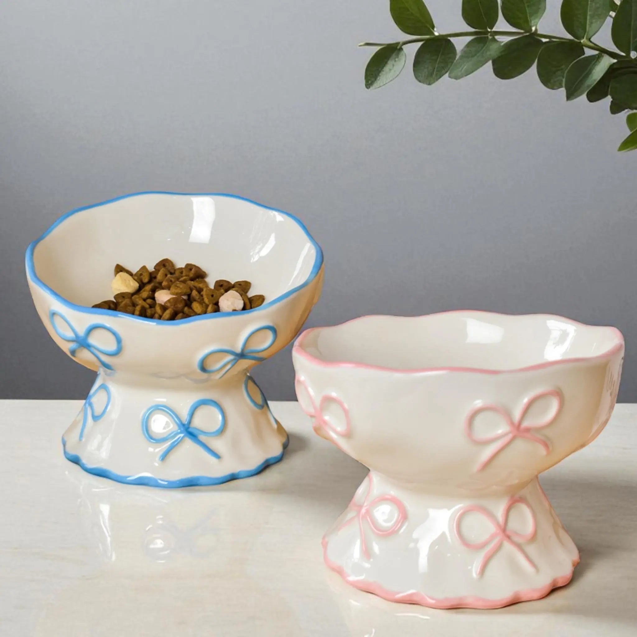 Two raised ceramic cat bowls with pink and blue ribbon patterns, one containing dry pet food on a white marble surface.
